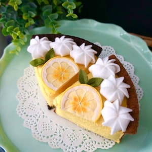 May include: Two slices of felt lemon meringue pie on a light blue plate with a white doily. The pie is decorated with white felt meringue and green felt leaves. The text "Divine Felt Studio" is visible in the bottom right corner of the image.