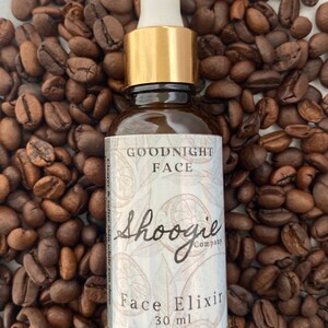 Puede incluir: Una botella de vidrio marrón con una tapa dorada y un gotero blanco. La botella está etiquetada con "Goodnight Face Shoogie Company Face Elixir 30 ml". La botella está rodeada de granos de café.