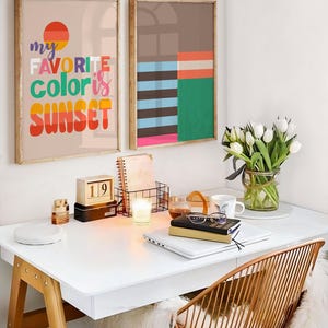 May include: A modern home office setup with two framed art prints, one with the text "my favorite color is sunset." A white desk holds a laptop, books, and a candle. A gold wire chair and a vase of white tulips complete the scene.