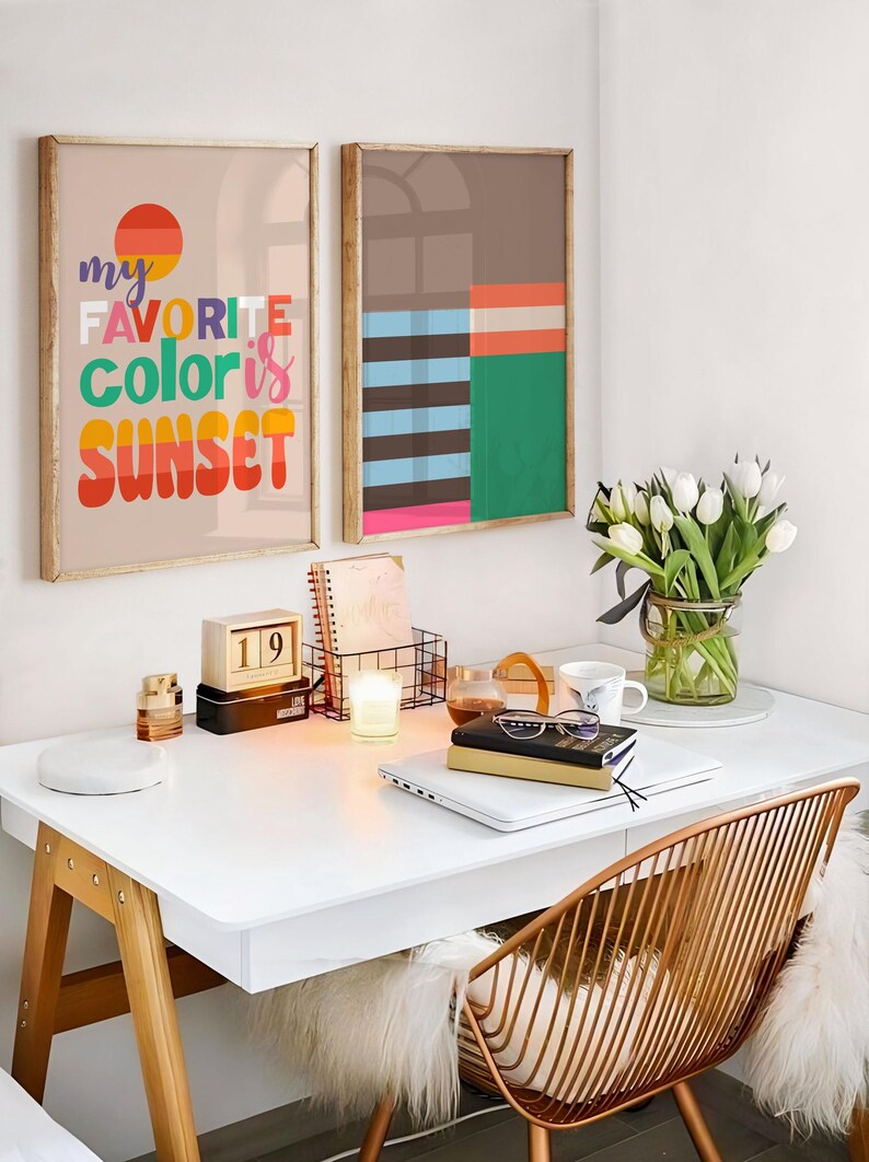 May include: A modern home office setup with two framed art prints, one with the text "my favorite color is sunset." A white desk holds a laptop, books, and a candle. A gold wire chair and a vase of white tulips complete the scene.