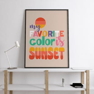 May include: A framed art print with the text "my FAVORITE color is SUNSET" in colorful, bold letters. The artwork features a stylized sunset graphic in shades of orange, yellow, and red, set against a neutral background. The print is displayed on a white shelf.