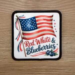 May include: Square coaster with a black border featuring a waving American flag design. The flag is red, white, and blue, with the text "Red, White & Blueberries" in a decorative font. A cluster of blueberries is at the bottom.