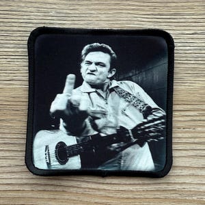 May include: Black and white patch featuring a portrait of a musician playing a guitar and giving the middle finger. The patch has a black border and is square-shaped. The musician is wearing a button-down shirt.