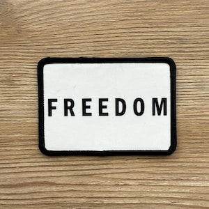 May include: A rectangular patch with the word "FREEDOM" in bold, black capital letters centered on a white background. The patch has a black border and is set against a light brown wooden surface.
