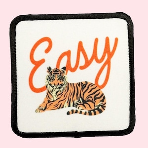 May include: A white square patch with a black border. The patch features an orange tiger lying down and the word "Easy" written in orange cursive.