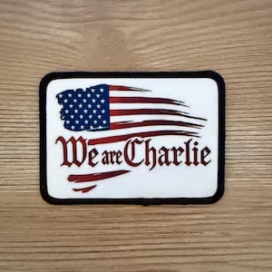 May include: Rectangular patch with a black border, featuring a stylized American flag design. The flag has blue stars and red and white stripes. The text "We are Charlie" is written in red script.