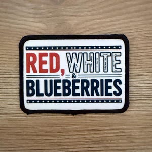 May include: A rectangular patch with the words "RED, WHITE & BLUEBERRIES" in red, white, and blue. The patch has a black border and is set against a wooden background.
