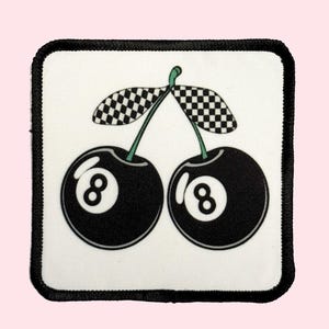 May include: A square patch featuring a whimsical design of two black 8-ball cherries with checkered leaves on a white background. The patch has a black border and is set against a light pink backdrop.