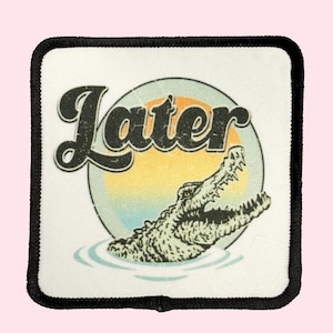 May include: A white patch with black stitching featuring a retro-style graphic of a crocodile emerging from water with a sunset behind it and the word "Later" in a black vintage font.
