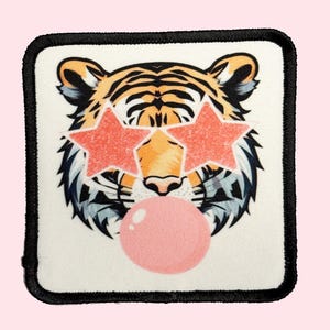 May include: A white square patch with a black border features a cartoon tiger wearing pink star-shaped sunglasses and blowing a pink bubble.