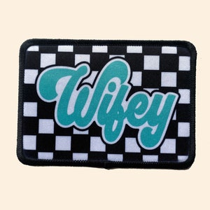 May include: A black and white checkered patch with the word "Wifey" in teal script.
