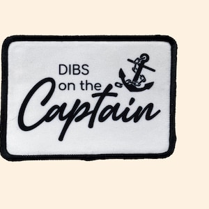 May include: White rectangular patch with a black border. The patch features the words "DIBS on the Captain" in a black, stylized font, along with a black anchor graphic. The patch is likely for clothing or accessories.
