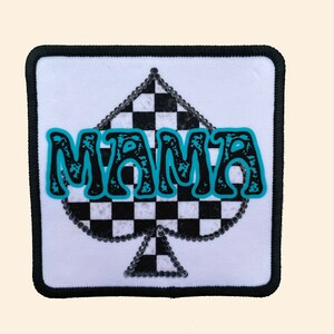 May include: A white patch with a black and white checkered spade design. The spade is outlined with silver rhinestones. The word "MAMA" is written in blue letters across the spade.