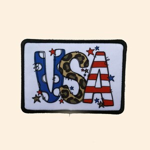 May include: A white patch with a black border featuring the word "USA" in colorful letters. The letters are decorated with polka dots, leopard print, and red and white stripes. There are small red stars scattered around the letters.