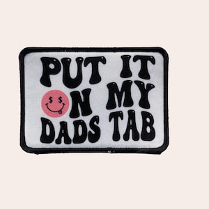 May include: White patch with black text that reads "Put it on my dads tab" with a pink smiley face with dollar signs for eyes.