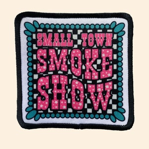 May include: A black and white checkered background with a pink and white text overlay that reads "Small Town Smoke Show". The text is surrounded by a teal blue and white border.