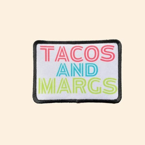 May include: A white rectangular patch with black stitching around the edges. The patch has the text "TACOS AND MARGS" in colorful, retro-style lettering.