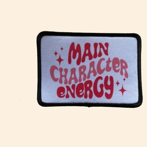 May include: White iron-on patch with black stitching around the edges. The patch features red text that reads "MAIN CHARACTER ENERGY" in a retro font with two small stars on either side of the text.