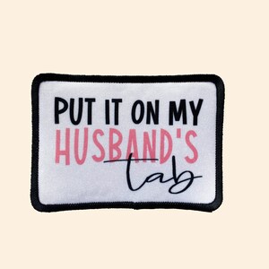 May include: White patch with black border and the text "PUT IT ON MY HUSBAND'S tab" in black and pink.