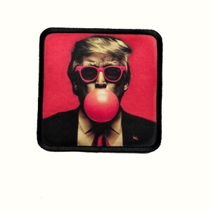 May include: A square patch with a black border and a red background. The image shows a man with blonde hair wearing pink sunglasses and blowing a pink bubble. The man is wearing a black suit and a red tie.