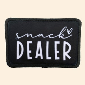 May include: Black rectangular patch with white text that reads "snack dealer" with a small heart symbol above the word "snack".