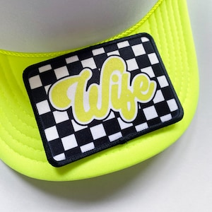 May include: A neon green trucker hat with a black and white checkered patch that says "Wife" in yellow script.