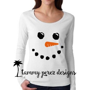 May include: White long-sleeved shirt with a black snowman face design. The snowman has black eyes, an orange carrot nose, and a black smile.