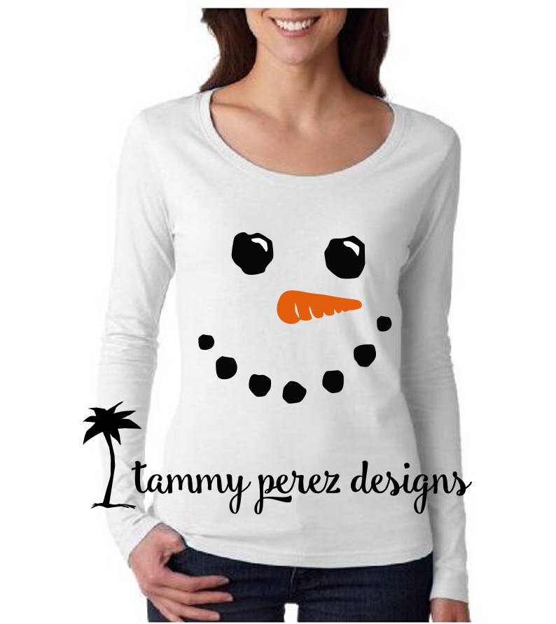 May include: White long-sleeved shirt with a black snowman face design. The snowman has black eyes, an orange carrot nose, and a black smile.