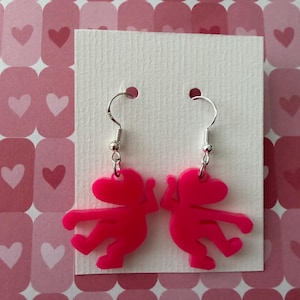 May include: A pair of bright pink earrings featuring a stylized figure with outstretched arms and legs. The earrings hang from silver-colored hooks. The background is a white card on a pink heart-patterned surface.