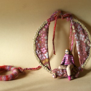 Può includere: Una bambola fatta a mano con abiti a righe rosa e bianche è seduta su un'altalena di tessuto floreale rosa e bianco. L'altalena è fatta di tessuto e filo ed è sospesa da una struttura in legno.