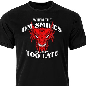 Op de afbeelding: Zwart T-shirt met een witte afbeelding van een rode draak met de tekst "When the DM Smiles It's Already Too Late"
