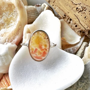Puede incluir: Un anillo de plata con una piedra ovalada de color rosa y amarillo engastada en un engaste de plata. El anillo está rodeado de conchas.