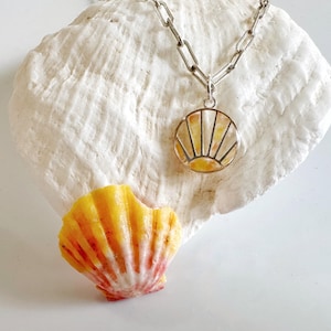 May include: A silver chain necklace with a round pendant featuring a sunrise design in yellow and orange. The necklace rests on a large, white seashell, with a smaller, yellow and red seashell nearby.