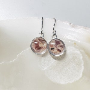 May include: A pair of silver-toned dangle earrings. Each earring features a circular, clear resin pendant with a mix of pink and beige organic inclusions. The earrings are displayed on a white, textured surface.