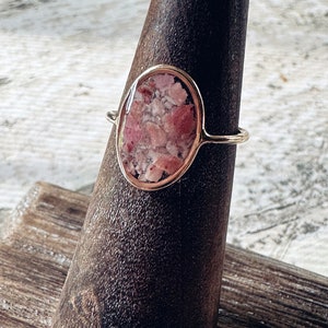 May include: A gold-colored ring with an oval-shaped, pink and white stone. The ring is set on a dark brown, wooden form. The stone appears to be made of small, irregular pieces.