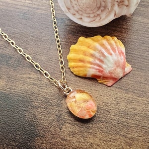 May include: A gold-colored chain necklace with an oval pendant. The pendant is filled with dried flowers in shades of orange and yellow. A seashell with orange and pink hues rests nearby on a wooden surface.