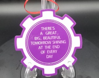 Könnte beinhalten: Ein lila-weißes zahnradförmiges Ornament mit dem Text "THERE'S A GREAT, BIG, BEAUTIFUL TOMORROW SHINING AT THE END OF EVERY DAY." Ein rotes Band ist zum Aufhängen befestigt. Das Ornament wird auf einem transparenten Ständer präsentiert.