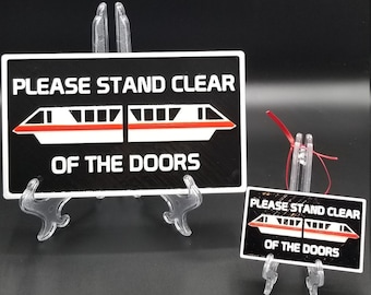 May include: Two rectangular black signs with white borders and white text that reads PLEASE STAND CLEAR OF THE DOORS. Each sign features a graphic of a white monorail train with a red stripe running horizontally across the centre of the train cars.