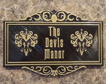 May include: A black and gold sign with the text "The Davis Manor" in gold lettering. The sign has a decorative border with a gold fleur-de-lis design on each side.