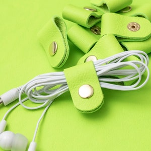 May include: A close-up of a set of lime green leather cord organisers with gold metal clasps. The organisers are holding a white cord with white earbuds.
