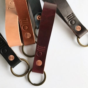 May include: Five leather keychains with metal rings. Each keychain is a different colour: brown, tan, black, dark brown, and burgundy. The keychains have the word "illo" embossed on them.