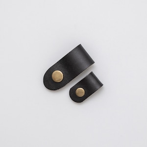 May include: Two black leather cord organisers with brass accents. The larger organiser is about 10 cm long and the smaller organiser is about 5 cm long.