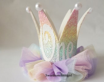 Op de afbeelding: Een regenboog glitterkroon met het woord "een" in witte glitter, versierd met witte parels en een veelkleurige tule strik.
