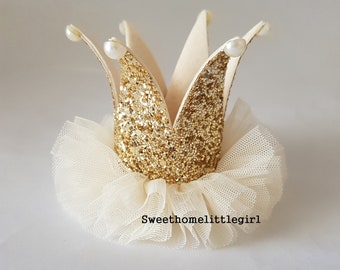May include: A gold glitter crown featuring a pointed design with small white faux pearls at each tip. The base of the crown is surrounded by a ruffled, off-white tulle fabric trim. The text "Sweethomelittlegirl" is printed in black below the crown.