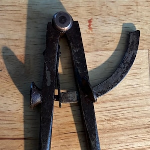 May include: An antique, rusty metal compass tool with two pointed legs and a curved arm. The tool has a central pivot and a small adjustment wheel. The compass is set against a wooden background.