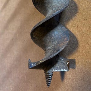 May include: Close-up of a vintage auger drill bit. The metal bit has a spiral design and a pointed tip. The tool is a dark, aged metal, showing signs of wear and use. The background is a neutral brown.