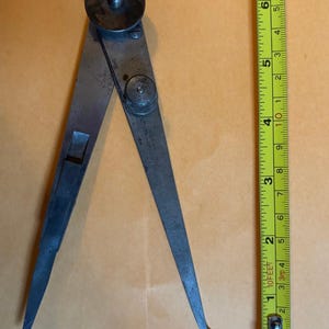 Puede incluir: Un calibrador de metal antiguo con puntas afiladas, utilizado para medir distancias. El calibrador está abierto, y una cinta métrica amarilla está al lado. La herramienta tiene una perilla de ajuste circular y está sobre una superficie marrón claro.