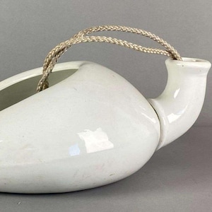 May include: A white ceramic bedpan with a curved spout and a handle made of braided rope. The bedpan has a wide, shallow basin and a smooth, glossy finish. The handle is attached to the top of the spout.