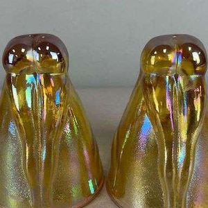 May include: A pair of bell-shaped salt and pepper shakers crafted from iridescent marigold carnival glass. Each shaker features a smooth, rounded top and a flared base with vertical molded ridges that catch the light with a metallic sheen.