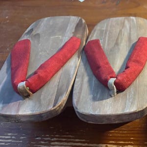 May include: Four pairs of traditional Japanese geta sandals. The wooden platforms have a natural finish, with fabric straps in red, blue-grey, and brown. The sandals are arranged in two rows, showcasing the different strap colors and styles.